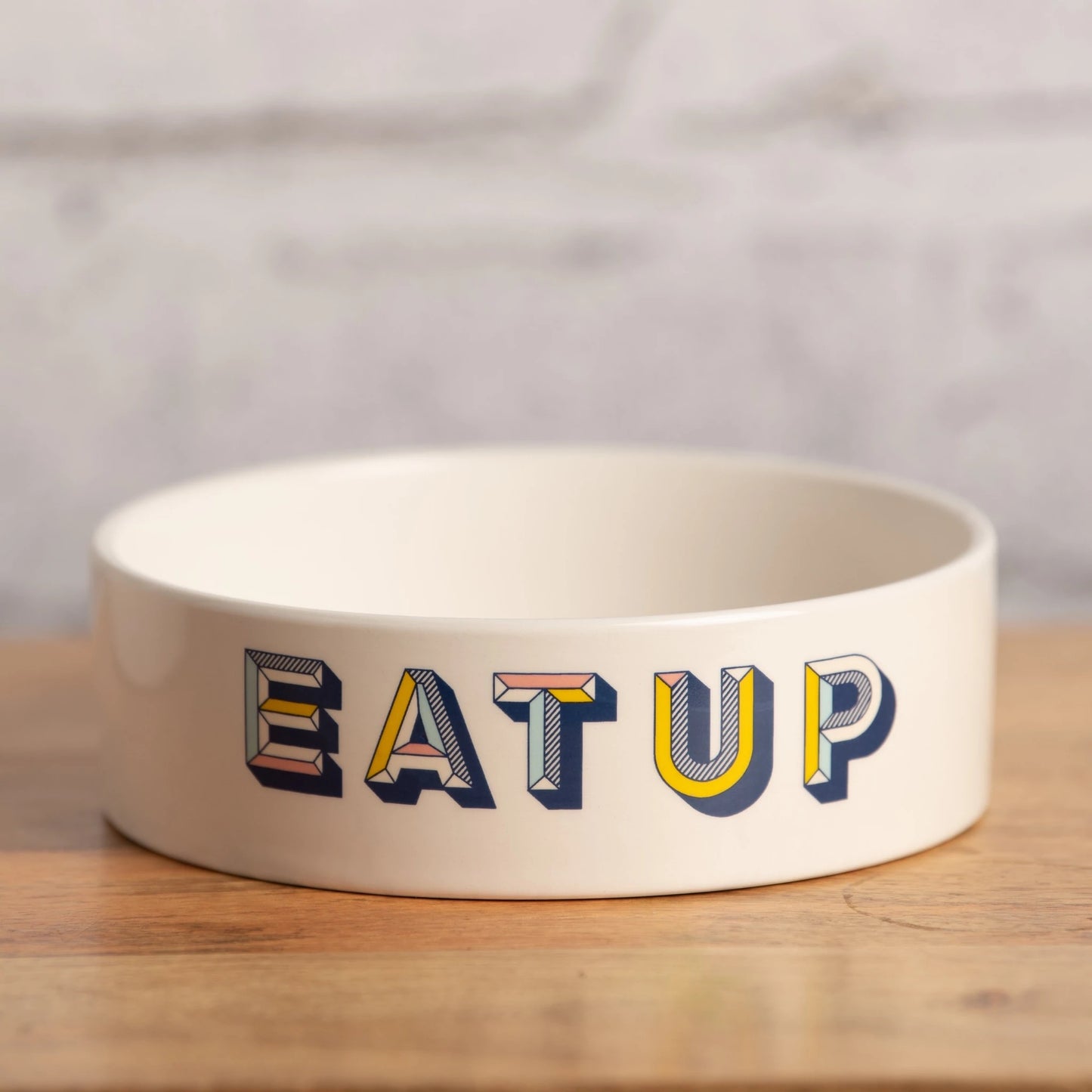 A beige ceramic pet bowl with a vibrant 'EAT UP' text design The bowl is round with a flat rim.