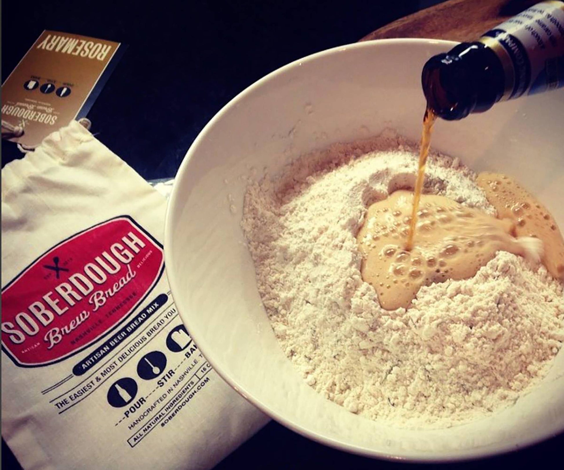 Bowl of flour with a bottle being poured into it, next to a bag labeled 'Soberdough Brew Bread'.
