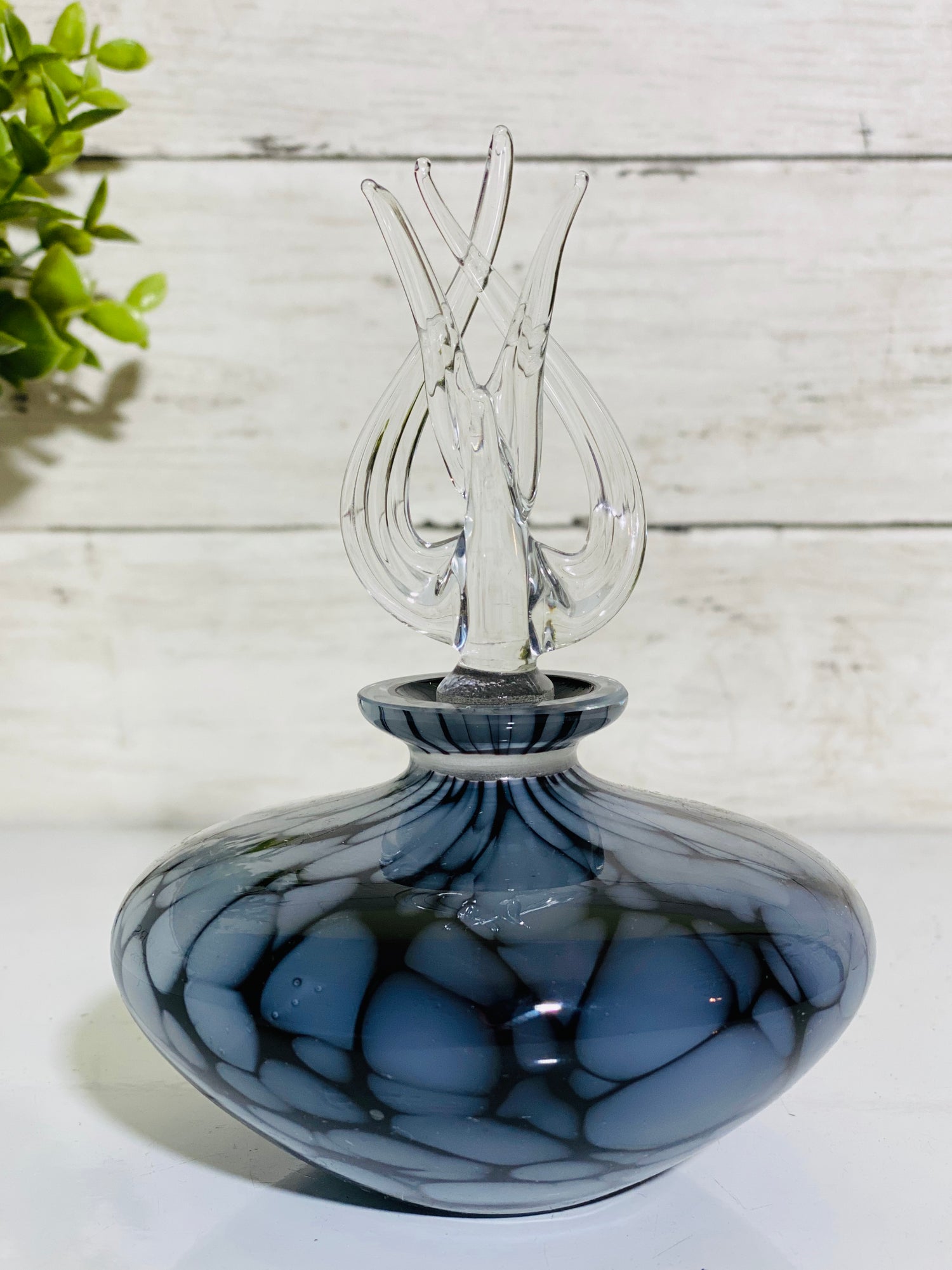 A flat, wide glass perfume bottle with a black and blue color scheme, featuring an intricate glass stopper.